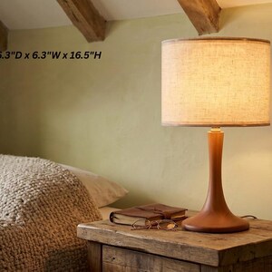 May include: A table lamp with a smooth, brown wooden base and a cylindrical, textured beige fabric shade. The lamp measures 6.3 inches deep, 6.3 inches wide, and 16.5 inches high, emitting a warm, soft glow from within the shade.