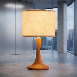 May include: A table lamp featuring a smooth, light-toned wooden base with a flared, circular pedestal. The lamp is topped with a cylindrical, textured beige fabric lampshade that emits a warm, soft glow, illuminating the surrounding space.