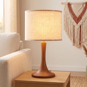May include: A table lamp with a smooth, hourglass-shaped wooden base and a cylindrical, off-white fabric lampshade. The lamp emits a warm, soft glow, illuminating the textured surface of the shade while resting on a square wooden side table.