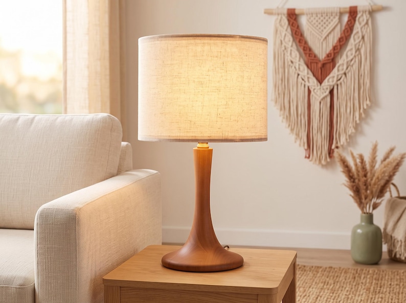 May include: A table lamp with a smooth, hourglass-shaped wooden base and a cylindrical, off-white fabric lampshade. The lamp emits a warm, soft glow, illuminating the textured surface of the shade while resting on a square wooden side table.