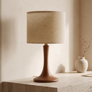 May include: A table lamp featuring a smooth, dark wood base with a curved, tapered stem and a wide, circular foundation. The lamp is topped with a cylindrical, textured beige fabric lampshade that diffuses light evenly across its surface.