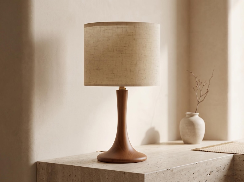 May include: A table lamp featuring a smooth, dark wood base with a curved, tapered stem and a wide, circular foundation. The lamp is topped with a cylindrical, textured beige fabric lampshade that diffuses light evenly across its surface.