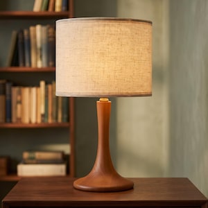 May include: A table lamp with a smooth, curved, medium-brown wooden base and a cylindrical, textured, off-white fabric lampshade. The lamp emits a warm, soft glow, illuminating the shade while resting on a flat, dark-toned wooden surface.