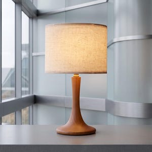 May include: A table lamp featuring a smooth, light brown wooden base with a curved, flared pedestal design. The lamp is topped with a cylindrical, textured off-white fabric lampshade that emits a soft, warm glow from the light source within.