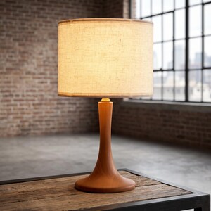 May include: A table lamp featuring a smooth, light brown wooden base with a curved, tapered stem and a wide, circular foundation. The lamp is topped with a cylindrical, off-white fabric lampshade that emits a warm, soft glow when illuminated.