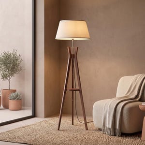 May include: A floor lamp with a dark brown wooden tripod base and a circular shelf, topped with a white fabric drum shade. The lamp features a slender central pole and a black power cord extending from the base to the floor.