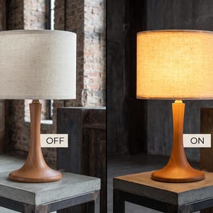 May include: A table lamp with a smooth, light brown wooden base and a cylindrical, textured off-white fabric shade. The image displays the lamp in two states: one labeled OFF with the light unlit, and one labeled ON with the light illuminated.