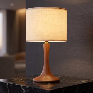 May include: A table lamp featuring a smooth, hourglass-shaped wooden base with a warm brown finish. The lamp is topped with a cylindrical, off-white fabric lampshade that emits a soft, diffused glow, creating a warm and inviting illumination.