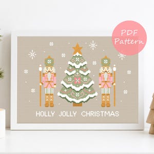 May include: A cross-stitch pattern in a white frame featuring a central green Christmas tree flanked by two pink nutcrackers. The design includes white snowflakes on a beige background with the text HOLLY JOLLY CHRISTMAS at the bottom.