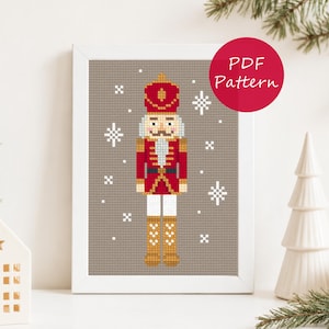 Può includere: Uno schema a punto croce in una cornice bianca raffigurante un soldatino schiaccianoci in uniforme rossa con dettagli dorati e un cappello alto, su sfondo tortora con fiocchi di neve bianchi e il testo PDF Pattern in un cerchio rosso.