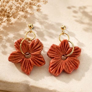 Peut inclure: Une paire de boucles d'oreilles pendantes avec des breloques en forme de fleur à cinq pétales dans une teinte orange terre cuite scintillante. Chaque fleur présente un motif texturé à rainures radiales, suspendue à un anneau doré.
