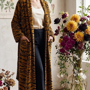 May include: A long, open-front coat featuring a mustard yellow and black tiger stripe pattern, worn over a cream-coloured camisole and dark navy blue trousers. The coat has long sleeves and two large patch pockets on the front.