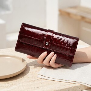 May include: A rectangular wallet in a deep burgundy color with a glossy, patent finish and a textured crocodile-skin pattern. The wallet features a flap closure secured by a central vertical strap with a small, square silver-toned metal accent.