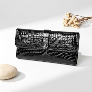 Puede incluir: Una cartera rectangular negra con acabado brillante y un patrón texturizado de piel de cocodrilo. La cartera cuenta con una solapa frontal asegurada por una correa vertical con un pequeño detalle de tachuela metálica en el centro.