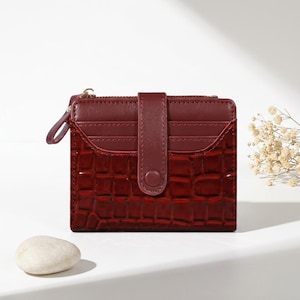 Peut inclure: Un portefeuille bifold rouge foncé présentant une texture gaufrée façon crocodile brillante sur la partie inférieure et une finition lisse sur la zone supérieure des fentes pour cartes. Il possède une patte de fermeture à bouton-pression.