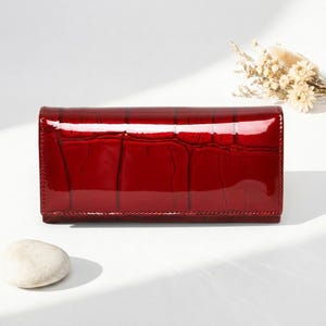 May include: A rectangular wallet in a deep, glossy red finish with a textured crocodile skin pattern. The wallet features a smooth, reflective surface and a clean, structured design with a horizontal orientation and visible stitching along the edges.