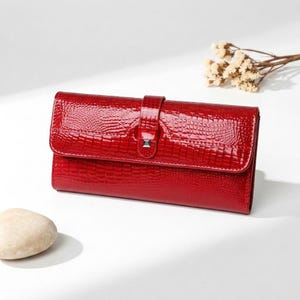 May include: A rectangular red wallet with a glossy finish and a textured crocodile skin pattern. The wallet features a front flap closure secured by a vertical strap with a small, square silver-toned metal accent piece centred on the front.