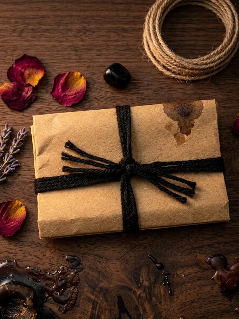 May include: A rectangular package wrapped in brown kraft paper, tied with black twine in a cross pattern. The paper features a dark brown stain on the upper right corner. The package is surrounded by dried rose petals, lavender sprigs, a black stone, and a coil of jute rope.