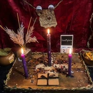 May include: A wooden board holds three lit purple candles, scattered dried herbs, and a small brown envelope tied with black string. A glass vase with dried grasses, a small bowl of dried petals, and a calendar block displaying Oct 23 sit nearby.