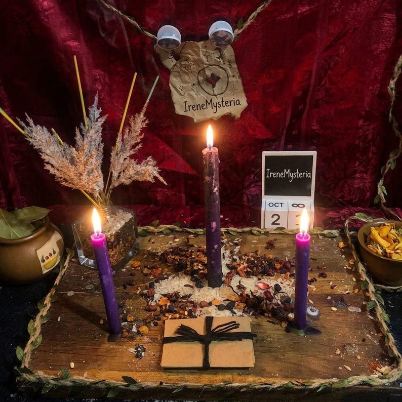 May include: A wooden board holds three lit purple candles, scattered dried herbs, and a small brown envelope tied with black string. A glass vase with dried grasses, a small bowl of dried petals, and a calendar block displaying Oct 23 sit nearby.