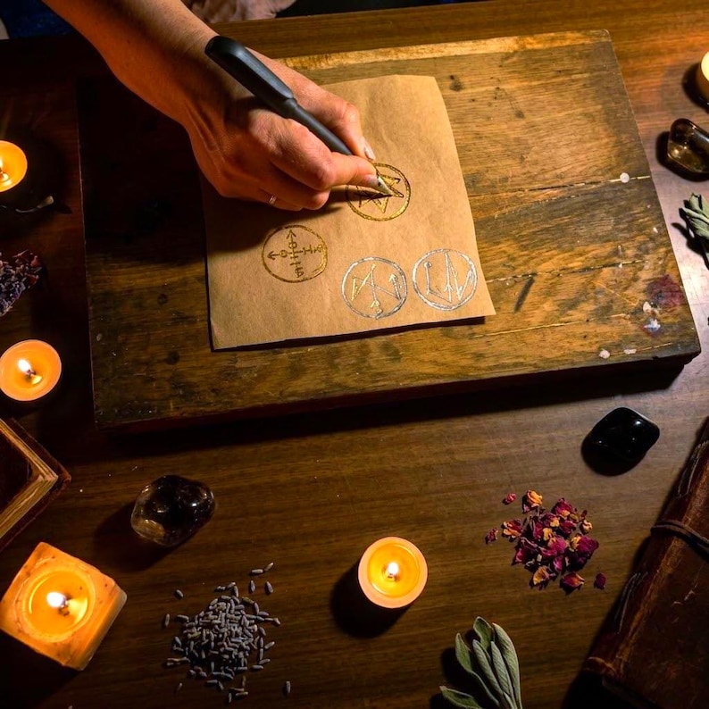May include: A hand uses a black stylus to draw circular symbols on a rectangular sheet of brown paper. The paper features gold and silver ink designs. The scene includes lit candles, scattered dried flower petals, and small piles of dried herbs.