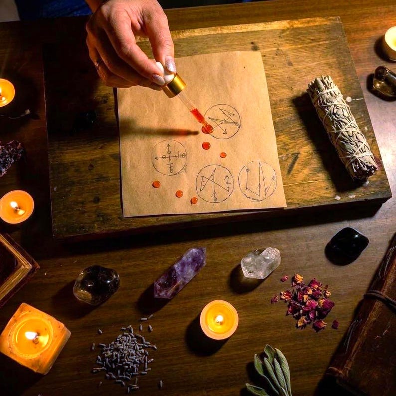 May include: A piece of tan paper with four hand-drawn circular symbols and scattered orange liquid droplets from a glass dropper. The scene includes a sage bundle, various crystals, dried rose petals, lavender, and several lit tea light candles.