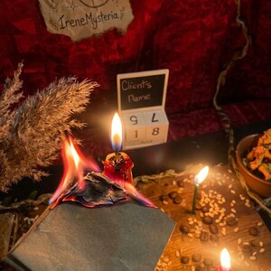 May include: A burning gray paper square with visible flames, positioned near a red candle, a small wooden block calendar displaying July 18, and a piece of parchment paper with the text IreneMysteria. Coffee beans and grains are scattered on the surface.