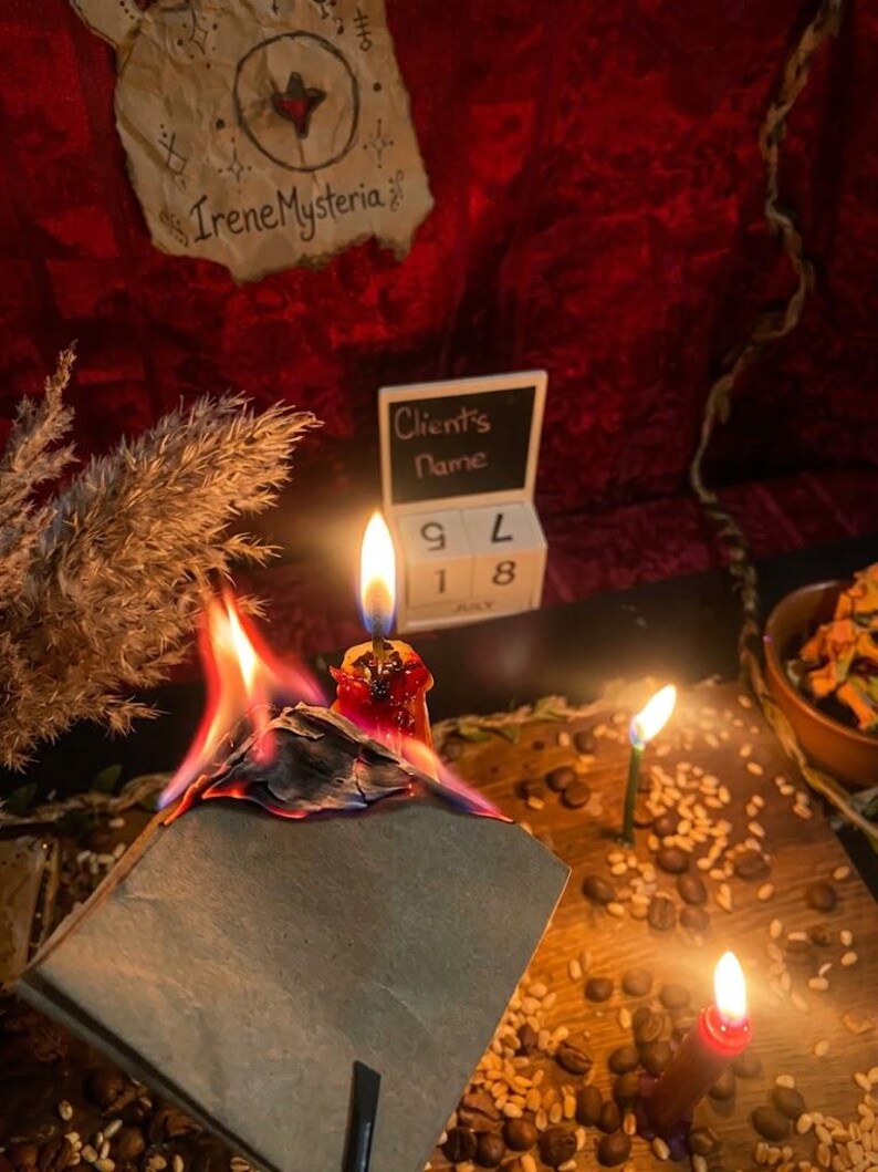 May include: A burning gray paper square with visible flames, positioned near a red candle, a small wooden block calendar displaying July 18, and a piece of parchment paper with the text IreneMysteria. Coffee beans and grains are scattered on the surface.