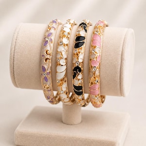 Może przedstawiać: Cztery bransoletki typu bangle w kolorze złotym z misternymi wzorami filigranowymi wypełnionymi emalią w kolorach fioletowym, białym, czarnym i różowym. Każda bransoletka jest ozdobiona małymi, przezroczystymi, okrągłymi kryształkami w metalowej oprawie.