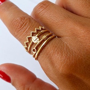 May include: A set of four stacked gold rings worn on a finger. The top ring features a zigzag crown design, the second ring has a small teardrop-shaped charm, the third ring has a textured twisted band, and the bottom ring is a smooth gold band.