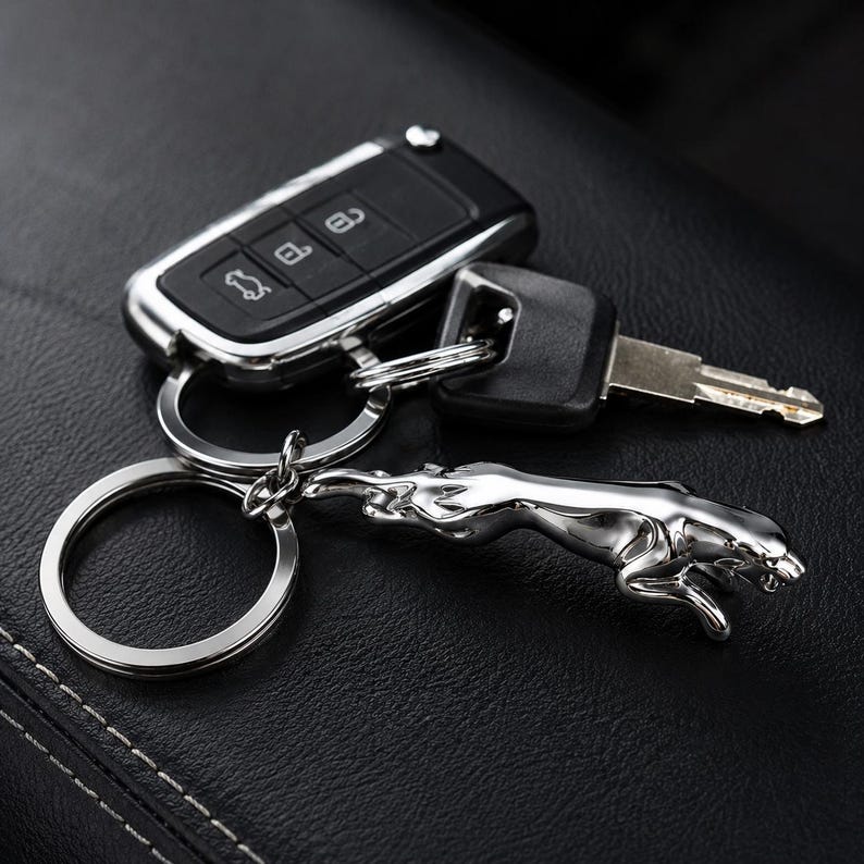 May include: A silver-toned metal keychain featuring a leaping jaguar figurine attached to a circular ring. The keychain is connected to a black plastic car key fob with three buttons and a metal key blade, all linked by a silver metal split ring.