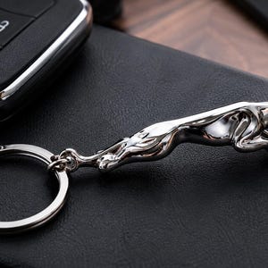 May include: A silver-toned metal keychain featuring a three-dimensional leaping jaguar figure attached to a circular split ring. The jaguar is depicted in a mid-leap pose with its body elongated and head forward, showcasing a polished, reflective finish.