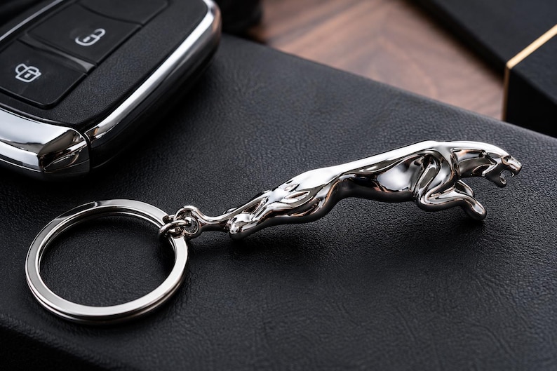May include: A silver-toned metal keychain featuring a three-dimensional leaping jaguar figure attached to a circular split ring. The jaguar is depicted in a mid-leap pose with its body elongated and head forward, showcasing a polished, reflective finish.