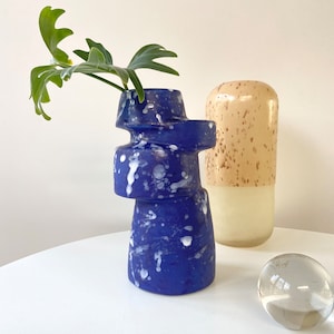 Op de afbeelding: Een blauwe en witte keramische vaas met een uniek, abstract ontwerp. De vaas is gemaakt van meerdere stukken die op elkaar zijn gestapeld. De vaas staat op een witte tafel naast een beige keramische vaas en een heldere glazen bol.