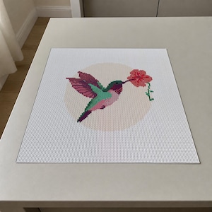 May include: A square cross-stitch embroidery project on white fabric featuring a hummingbird with green, pink, and purple plumage hovering near a red flower. The design is centered within a pale circular background, showcasing detailed needlework.