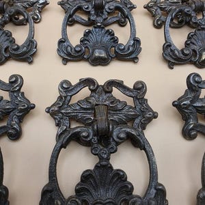 May include: A collection of dark grey metal drawer pulls featuring an ornate, symmetrical design with intricate scrollwork and a central drop handle. Each piece has a textured, weathered finish with raised floral-inspired decorative elements.