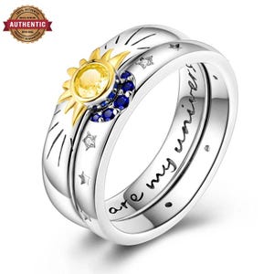 May include: A silver ring featuring a yellow gemstone sun and blue gemstone crescent moon motif. The band is engraved with the text 'you are my universe' and includes small star-shaped cutouts. The design incorporates sunburst rays.