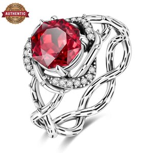 Könnte beinhalten: Ein Silberring mit einem großen, facettierten, achteckigen roten Edelstein in der Mitte, umgeben von einem Kranz aus kleinen, klaren, rund geschliffenen Steinen. Die Ringschiene hat ein strukturiertes, ineinander verschlungenes Rankenmuster.