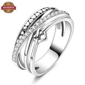 Puede incluir: Un anillo de plata con un diseño cruzado de varias bandas. Una banda está adornada con una hilera de pequeñas piedras transparentes, mientras que otra muestra un patrón repetitivo de pequeños corazones. El interior tiene grabado el texto 925.