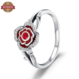 Puede incluir: Un anillo de plata con un motivo central de rosa, pétalos de esmalte rojo y una piedra roja en el centro. La rosa está bordeada por pequeñas piedras claras y el aro tiene un diseño de tallo con espinas, con el sello 925 en el interior.