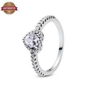 May include: A silver ring featuring a central heart-shaped clear stone surrounded by a halo of smaller round clear stones. The band is also adorned with a row of small round clear stones extending along the top half of the metal shank.