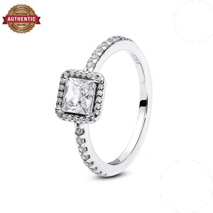 Op de afbeelding: Een zilveren ring met een vierkant geslepen heldere steen in het midden, omringd door een halo van kleine ronde heldere steentjes. De ringband is aan de bovenste helft ook bezet met kleine ronde heldere steentjes. De binnenkant is gestempeld met S925.