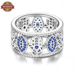 May include: A silver band ring featuring an alternating pattern of openwork hamsa hand motifs and marquise-shaped eye symbols. The eye symbols are inlaid with small, vibrant blue stones, and the ring edges are detailed with a beaded texture.