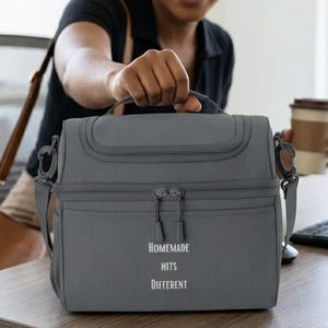 Puede incluir: Una bolsa de almuerzo aislada de color gris carbón con un asa superior y una correa de hombro desmontable. El frente presenta un bolsillo con cremallera y el texto impreso en blanco "HOMEMADE HITS DIFFERENT". Tiene una forma rectangular.