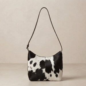 May include: A shoulder bag featuring a black and white cowhide pattern with a textured fur finish. The bag has a curved top silhouette, a single thin dark brown leather strap with an adjustable buckle, and a central vertical seam on the front.