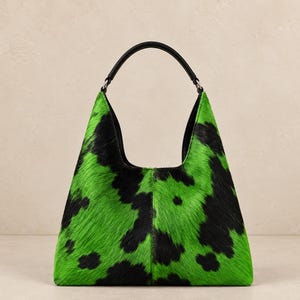 May include: A hobo-style handbag featuring a vibrant neon green and black cow-print pattern with a textured hair-on-hide finish. The bag has a single black leather handle attached with silver-toned metal hardware and a structured base.