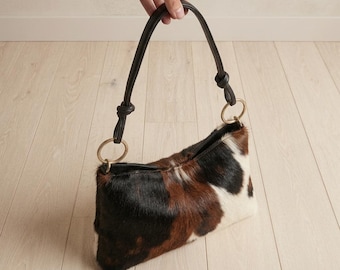 Könnte beinhalten: Eine kleine Handtasche mit einem dreifarbigen Kuhfellmuster in Schwarz, Braun und Weiß. Die Tasche hat einen einzelnen schwarzen Lederriemen, der an beiden Seiten an goldfarbenen Metallringen befestigt ist, mit Knoten an den Verbindungspunkten.