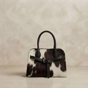 Könnte beinhalten: Eine Handtasche mit schwarz-weißem Kuhfellmuster und strukturierter Felloberfläche. Die Tasche verfügt über zwei schwarze Ledergriffe und einen horizontalen schwarzen Lederriemen auf der Vorderseite mit einer silberfarbenen Metallschnalle.