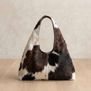 Puede incluir: Un bolso de hombro estilo hobo confeccionado en piel de vaca con un distintivo patrón tricolor de manchas blancas, marrón oscuro y negras. El bolso tiene un asa integrada única y una costura vertical que recorre el centro del panel frontal.