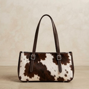 Pode incluir: Uma bolsa retangular com um padrão de pele de vaca castanho e branco no painel frontal. A bolsa inclui duas alças de ombro em couro castanho escuro presas com fivelas de metal prateado e um acabamento em couro castanho escuro.